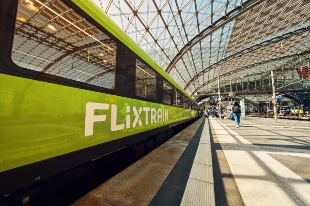 Ein grüner FlixTrain steht an einem Bahnsteig in einem modernen, lichtdurchfluteten Bahnhof mit gläsernem Dach. Menschen laufen entlang des Bahnsteigs, einige mit Gepäck. Die Perspektive zeigt den Zug in Nahaufnahme auf der linken Seite, während sich das Glasdach bogenförmig über die gesamte Szene spannt.