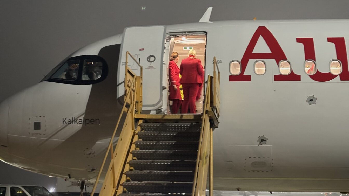 Ein Flugzeug der Austrian Airlines steht bei Nacht auf dem Rollfeld des Flughafens. Drei Crewmitglieder in roten Uniformen stehen in der geöffneten Flugzeugtür und unterhalten sich. Eine mobile Treppe führt zur Tür hinauf. Der Flugzeugname „Kalkalpen“ ist neben dem Cockpit sichtbar, wo zwei Pilot*innen sitzen. Die Szene ist leicht nebelig beleuchtet.