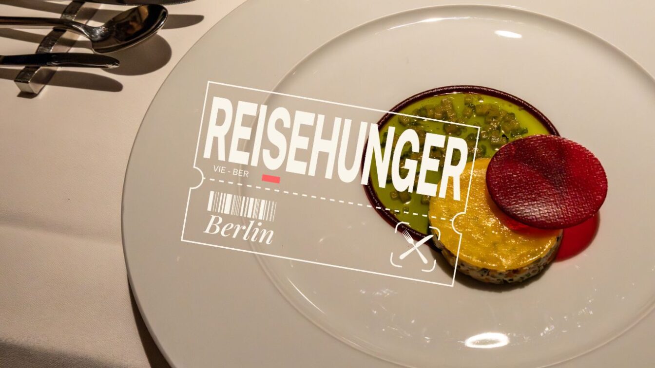 Ein edel angerichteter Teller mit einem kunstvollen veganen Gericht: grüne und gelbe Gemüsekomponenten, garniert mit einer runden, knallroten Scheibe – vermutlich Rote Bete – auf einem weißen Porzellanteller. Links im Bild glänzendes Besteck auf weißer Tischdecke. Darüber liegt ein grafisches Overlay mit dem Schriftzug „REISEHUNGER“, einem stilisierten Flugticket von Wien nach Berlin (VIE – BER), darunter die Wortmarke „Berlin“ und ein kleiner Flugzeug-Icon. Das Bild vermittelt kulinarische Reiselust mit Fokus auf Fine Dining in Berlin.