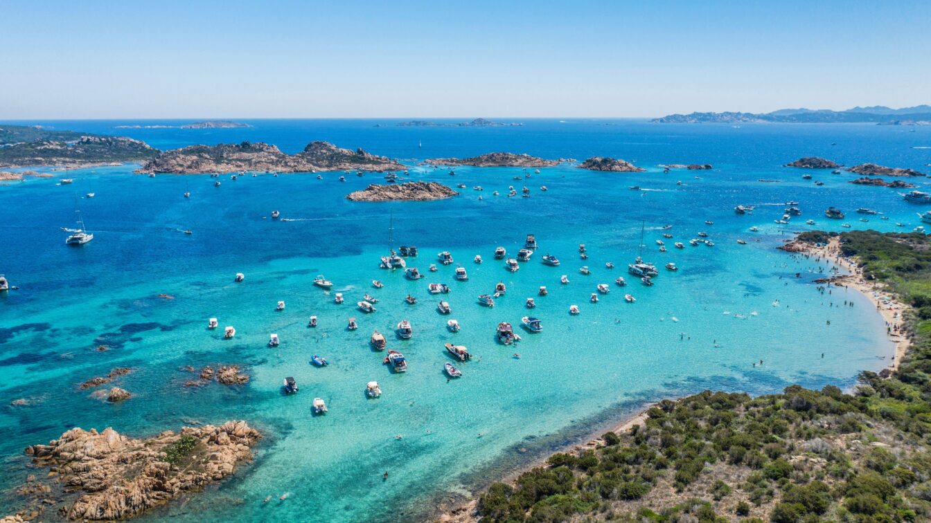 Luftaufnahme vom türkisblauen Meer vor Sardinien mit zahlreichen Booten vor der Küste und feinem Sandstrand – beliebtes Sommerziel in Italien mit kristallklarem Wasser und mediterraner Landschaft.