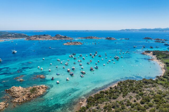 Luftaufnahme vom türkisblauen Meer vor Sardinien mit zahlreichen Booten vor der Küste und feinem Sandstrand – beliebtes Sommerziel in Italien mit kristallklarem Wasser und mediterraner Landschaft.