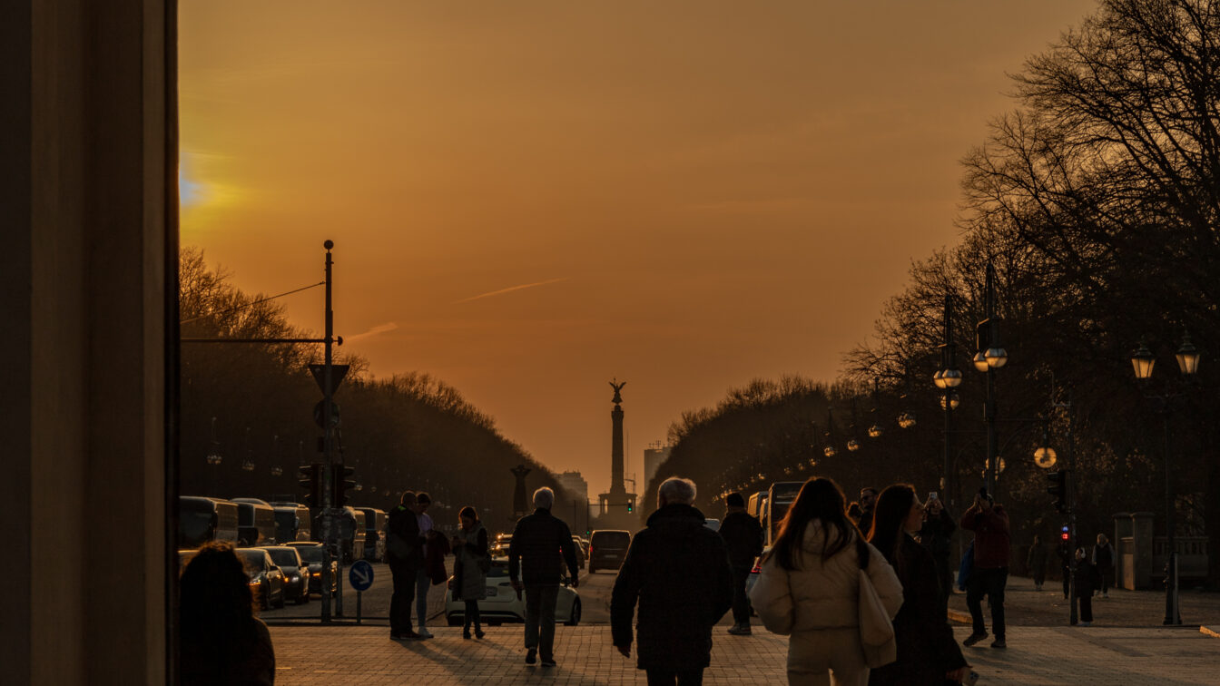 Menschen spazieren bei Sonnenuntergang auf der Straße des 17. Juni in Berlin, mit Blick auf die goldene Siegessäule in der Ferne. Die tiefstehende Sonne taucht den Himmel in warme Orangetöne, während Laternenlichter entlang der Straße leuchten. Bäume säumen den Weg, Silhouetten und Schatten betonen die ruhige Abendstimmung.