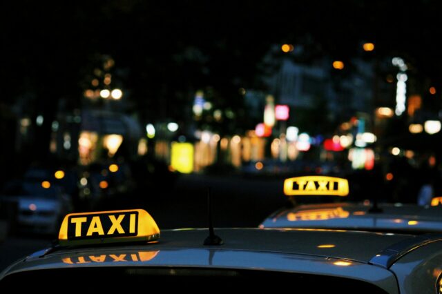 Zwei beleuchtete Taxi-Schilder auf den Dächern weißer Autos in einer nächtlichen, unscharf beleuchteten Stadtstraße. Im Hintergrund leuchten verschwommene Lichtquellen von Geschäften, Autos und Straßenlaternen.
