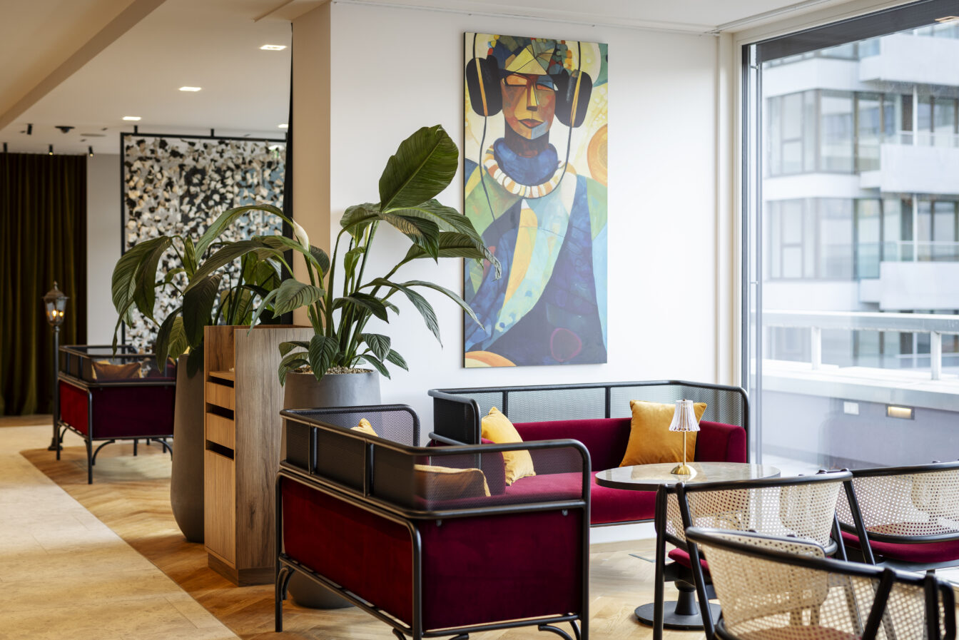 Stilvoller Loungebereich im ARCOTEL Wien mit samtroten Sofas, gelben Zierkissen und runden Marmortischen. An der Wand hängt ein farbenfrohes Gemälde einer abstrahierten Person mit Kopfhörern. Große grüne Pflanzen in modernen Töpfen trennen die Sitznischen voneinander. Durch das Fenster fällt viel Tageslicht.