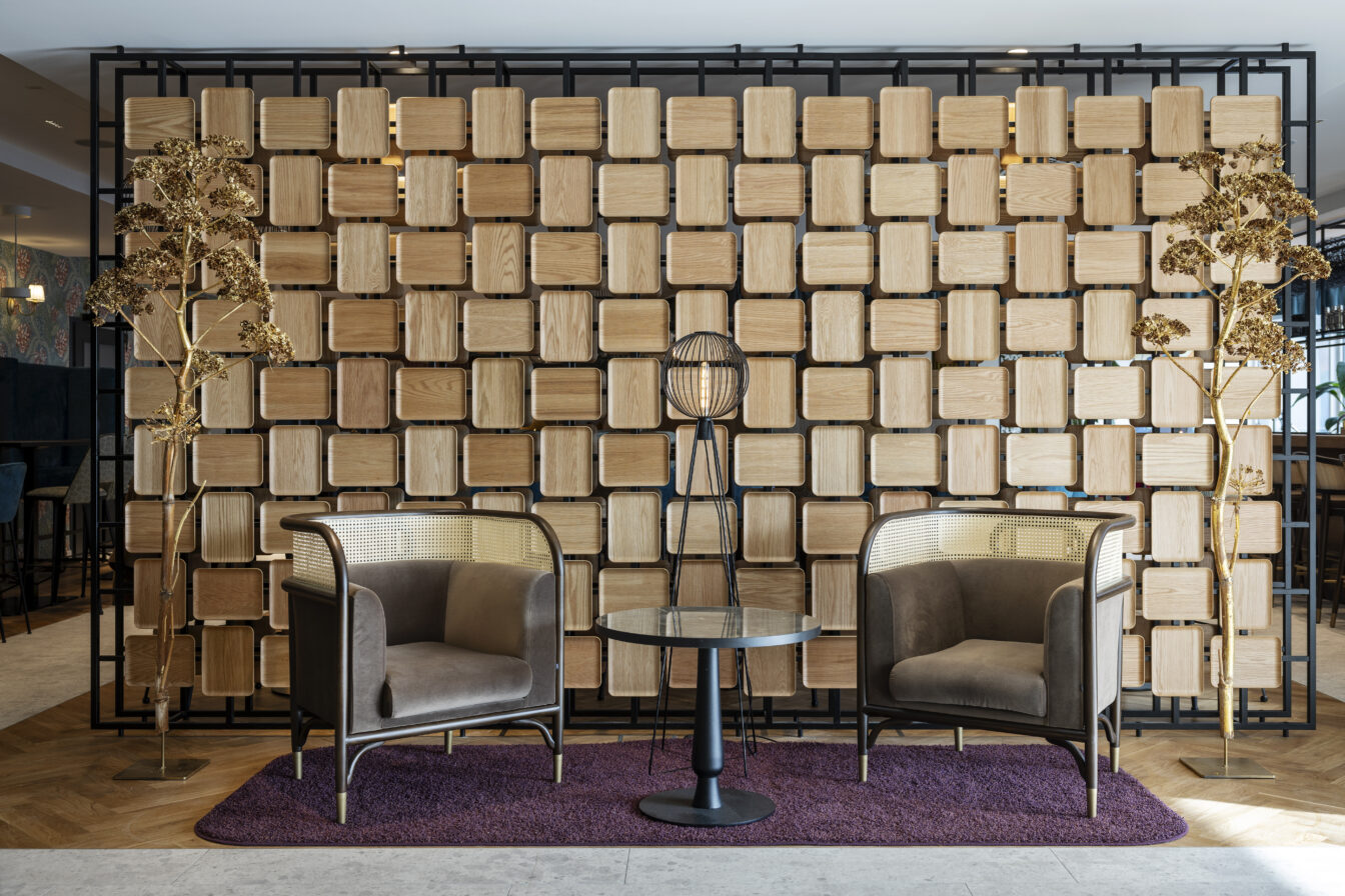 Zwei gemütliche, braune Sessel mit Wiener Geflecht stehen einander gegenüber auf einem lila Teppich im ARCOTEL Wien. Dazwischen ein kleiner runder Tisch und eine moderne Stehlampe mit Gitter-Lampenschirm. Im Hintergrund eine dekorative Raumtrennung aus quadratischen Holzelementen und goldfarbenen Trockenpflanzen.