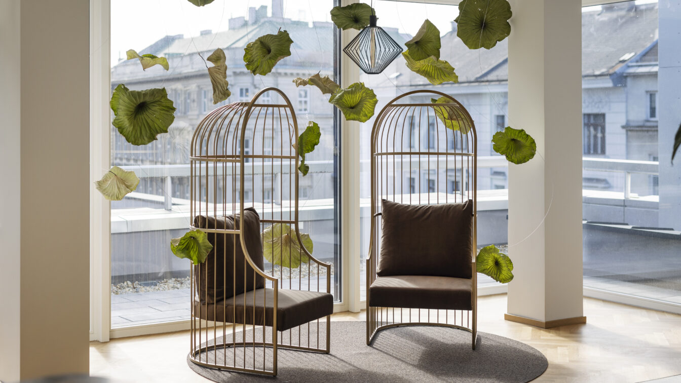 Zwei moderne, vogelkäfigartige Sessel mit braunen Polstern stehen einander gegenüber auf einem runden Teppich in einem lichtdurchfluteten Raum mit Panoramafenstern im ARCOTEL Wien. Zwischen den Sesseln schweben dekorative grüne Blätter, die an dünnen Fäden von der Decke hängen. Im Hintergrund ist die Wiener Innenstadt zu sehen