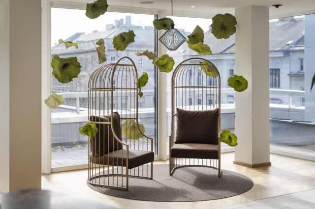 Zwei moderne, vogelkäfigartige Sessel mit braunen Polstern stehen einander gegenüber auf einem runden Teppich in einem lichtdurchfluteten Raum mit Panoramafenstern im ARCOTEL Wien. Zwischen den Sesseln schweben dekorative grüne Blätter, die an dünnen Fäden von der Decke hängen. Im Hintergrund ist die Wiener Innenstadt zu sehen