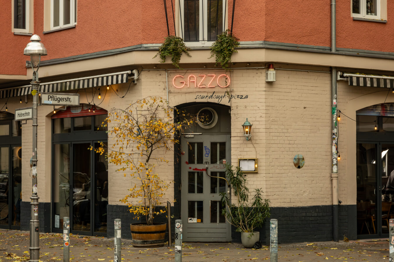 Eingang zur Pizzeria „Gazzo“ an der Ecke Pflügerstraße und Hobrechtstraße in Berlin-Neukölln. Die Fassade ist hell gestrichen, darüber ein Neon-Schriftzug „GAZZO“ mit dem Zusatz „sourdough pizza“. Vor der Tür stehen zwei große Pflanzen in Töpfen, daneben ein kleiner Baum mit gelben Herbstblättern. Gemütliche Atmosphäre mit Lichterkette über dem Eingang und Blick ins Innere des Lokals.