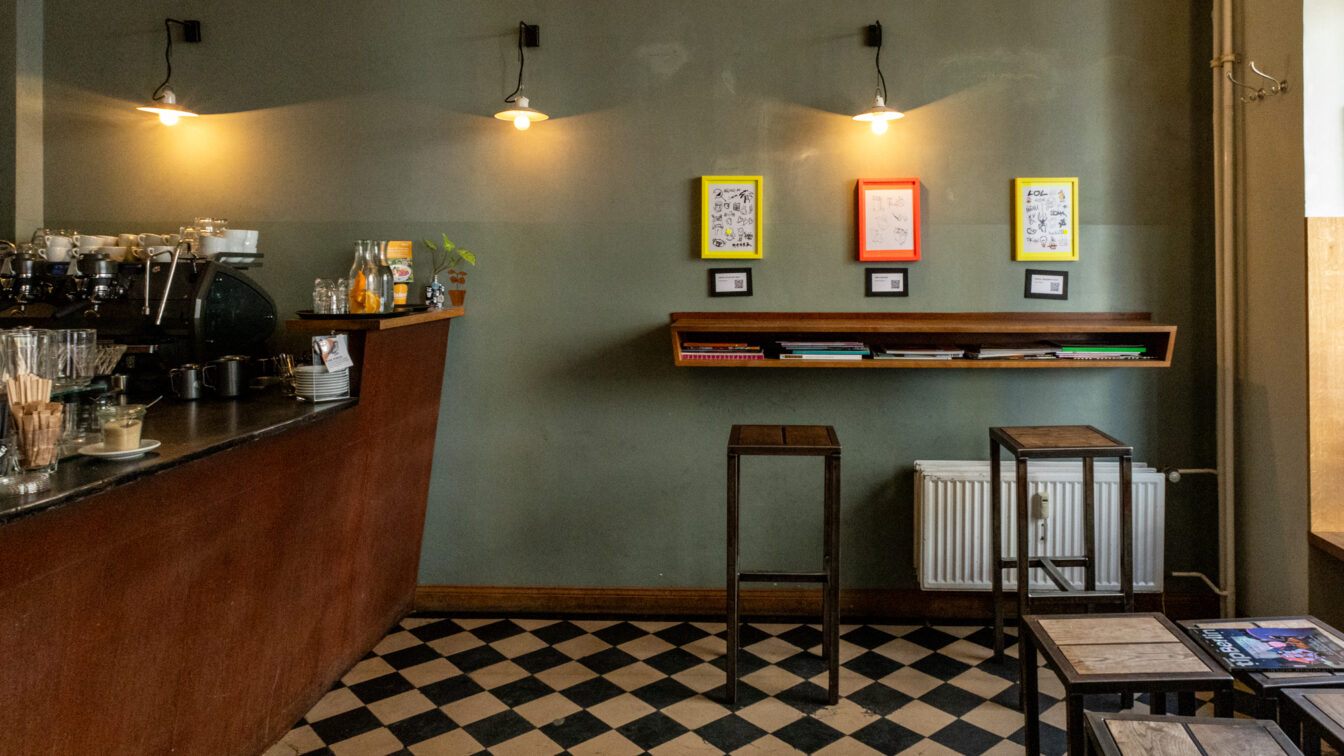 Gemütliches Café mit dunklem Thekenbereich links, Espressomaschine, Gläsern und Getränken. Die Wand ist in einem matten Grünton gestrichen und mit drei farbigen Bilderrahmen (gelb, rot, gelb) dekoriert, darunter kleine QR-Code-Schilder. Darunter ein schräges Wandregal mit Magazinen und Büchern. Zwei schlichte Barhocker stehen davor. Schwarz-weiß karierter Boden, sanfte Beleuchtung durch drei Wandleuchten. Rechts im Bild einfache Holztische mit Magazinen. Ruhige, minimalistische Atmosphäre.