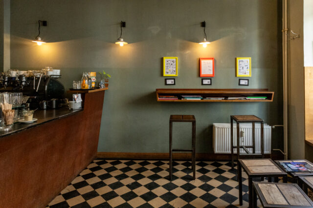 Gemütliches Café mit dunklem Thekenbereich links, Espressomaschine, Gläsern und Getränken. Die Wand ist in einem matten Grünton gestrichen und mit drei farbigen Bilderrahmen (gelb, rot, gelb) dekoriert, darunter kleine QR-Code-Schilder. Darunter ein schräges Wandregal mit Magazinen und Büchern. Zwei schlichte Barhocker stehen davor. Schwarz-weiß karierter Boden, sanfte Beleuchtung durch drei Wandleuchten. Rechts im Bild einfache Holztische mit Magazinen. Ruhige, minimalistische Atmosphäre.