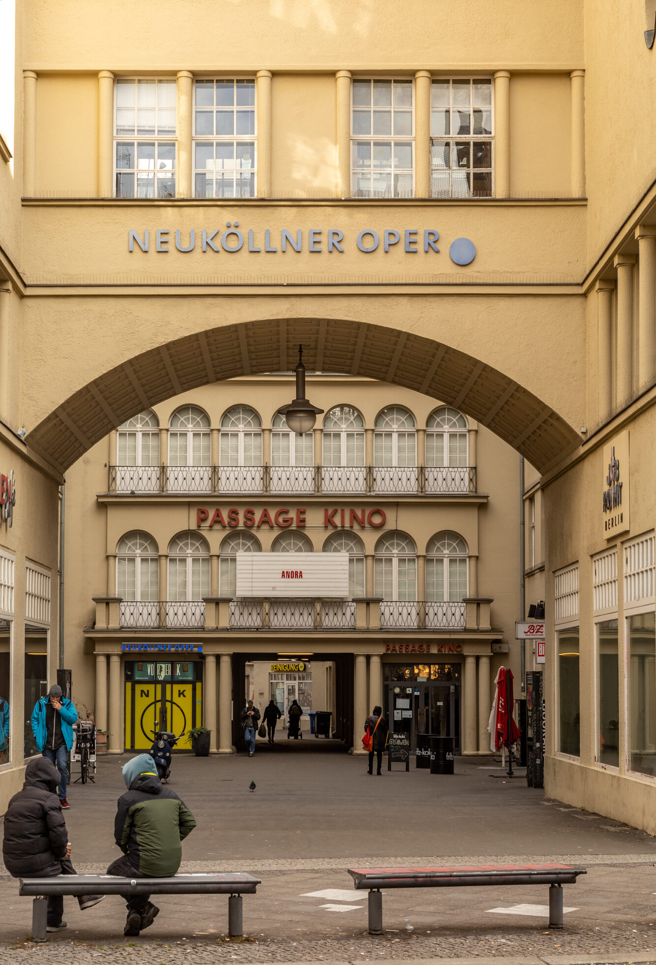 Blick auf den Eingangsbereich der „Neuköllner Oper“ und des „Passage Kinos“ in Berlin-Neukölln. Ein gelbes Gebäude mit großen Rundbogenfenstern und einem Durchgang, über dem in roten Buchstaben „PASSAGE KINO“ steht. Auf dem Leuchtschild der Filmname „Anora“. Menschen gehen oder sitzen auf Bänken im Vordergrund, teils in Winterkleidung. Die Szene wirkt ruhig und urban.