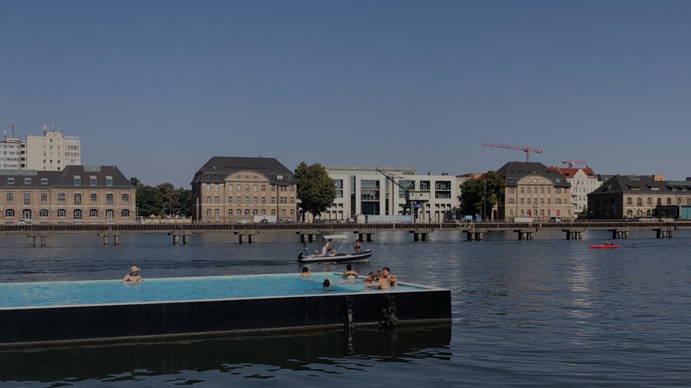 Sommerliche Szene im Badeschiff Berlin – ein schwimmendes Freibad in der Spree mit Blick auf historische Gebäude und moderne Architektur am gegenüberliegenden Ufer. Menschen genießen das erfrischende Wasser unter blauem Himmel – eines der außergewöhnlichsten Schwimmbäder in Berlin