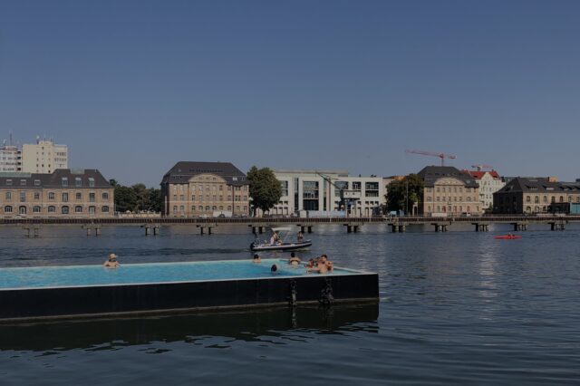 Sommerliche Szene im Badeschiff Berlin – ein schwimmendes Freibad in der Spree mit Blick auf historische Gebäude und moderne Architektur am gegenüberliegenden Ufer. Menschen genießen das erfrischende Wasser unter blauem Himmel – eines der außergewöhnlichsten Schwimmbäder in Berlin