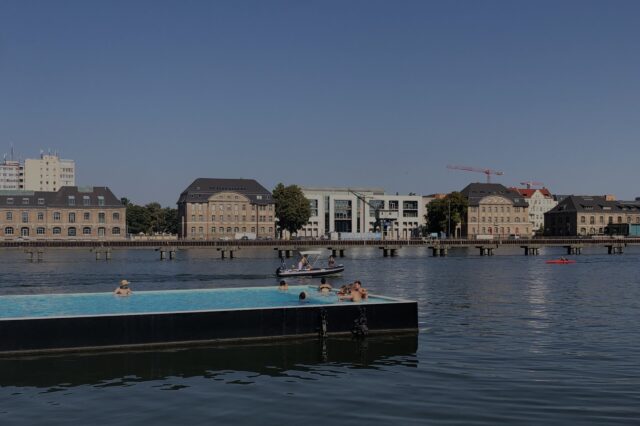 Sommerliche Szene im Badeschiff Berlin – ein schwimmendes Freibad in der Spree mit Blick auf historische Gebäude und moderne Architektur am gegenüberliegenden Ufer. Menschen genießen das erfrischende Wasser unter blauem Himmel – eines der außergewöhnlichsten Schwimmbäder in Berlin