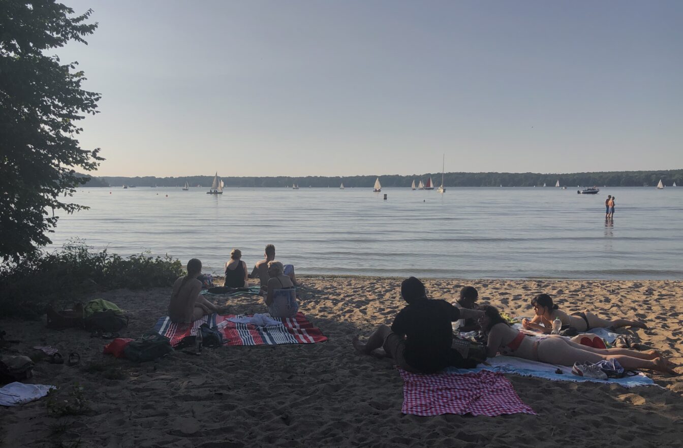 Menschen sitzen und liegen entspannt auf Picknickdecken am Sandstrand eines ruhigen Sees, während im Hintergrund zahlreiche Segelboote auf dem Wasser treiben. Die Szene spielt sich an einem sonnigen, warmen Tag ab, das Licht ist weich und sommerlich. Einige Personen stehen im flachen Wasser, andere genießen die Aussicht aufs Wasser. Links ragen Bäume ins Bild und spenden Schatten.