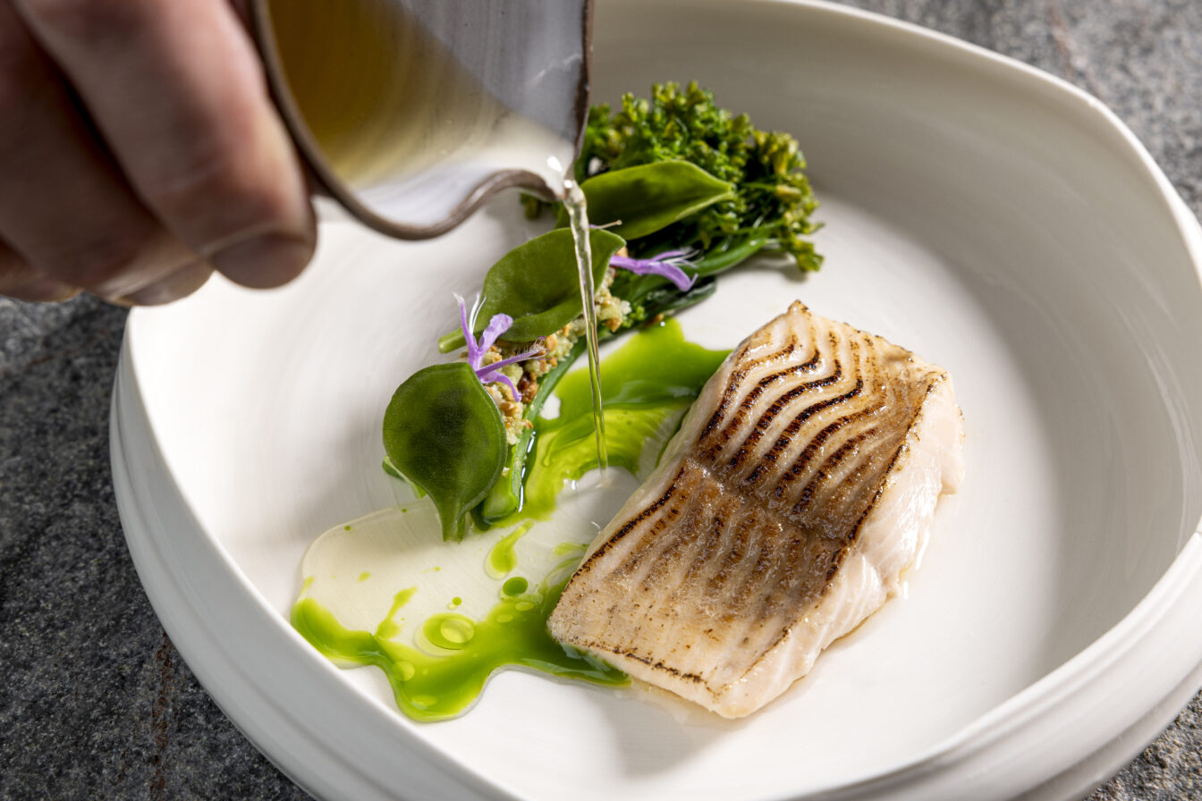 Gourmetgericht mit zart gebratener Forelle, grüner Kräutersauce, frischen Wildkräutern und essbaren Blüten – edle Fine-Dining-Komposition mit Sauce am Teller angerichtet, serviert auf handgefertigtem Porzellan.