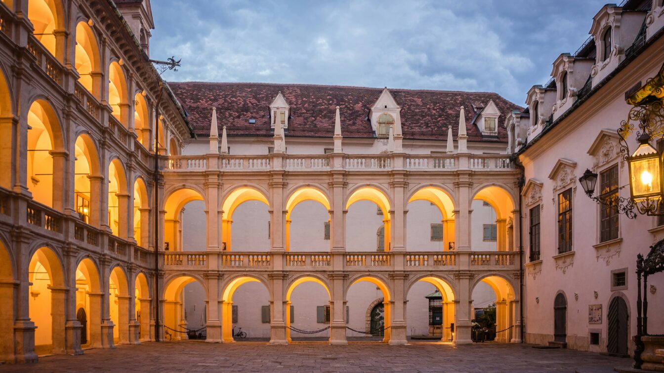 Innenhof der Grazer Burg im Abendlicht mit zwei Stockwerken offener Arkadengänge im Renaissancestil. Die warm beleuchteten Rundbögen und das Kopfsteinpflaster erzeugen eine stimmungsvolle Atmosphäre. Über dem Hof wölbt sich ein bewölkter Abendhimmel.