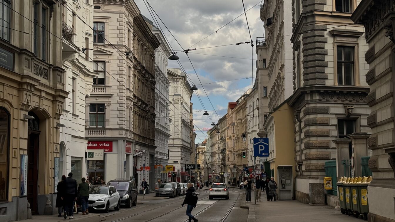 Straßenszene auf der Josefstäder Straße in Wien, Blickrichtung stadteinwärts. Historische Fassaden säumen die Straße, an der Straßenbahnschienen verlaufen. Menschen gehen am Gehsteig, Autos parken am Rand, ein paar fahren in der Mitte. Bewölkter Himmel mit Sonnenstrahlen, die durch die Wolken brechen.
