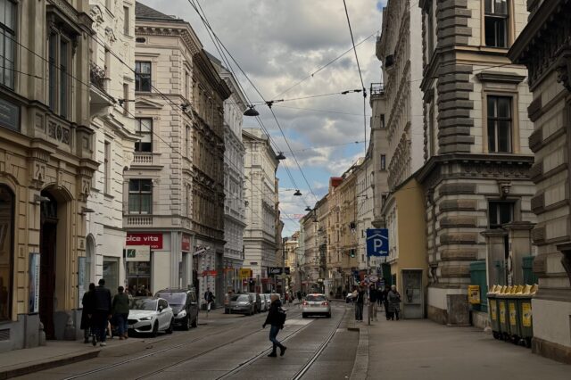 Straßenszene auf der Josefstäder Straße in Wien, Blickrichtung stadteinwärts. Historische Fassaden säumen die Straße, an der Straßenbahnschienen verlaufen. Menschen gehen am Gehsteig, Autos parken am Rand, ein paar fahren in der Mitte. Bewölkter Himmel mit Sonnenstrahlen, die durch die Wolken brechen.