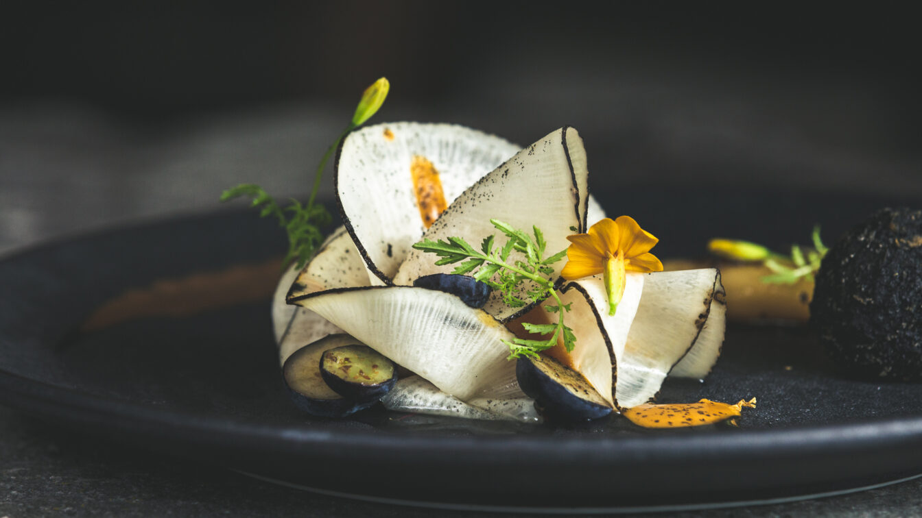 Gourmetgericht im Restaurant Kräuterreich by Vitus Winkler: kunstvoll angerichtete Komposition aus hauchdünnem Gemüse, Wildkräutern, Blüten und feinen Texturen auf dunklem Teller – moderne Alpenküche auf Sterne-Niveau.