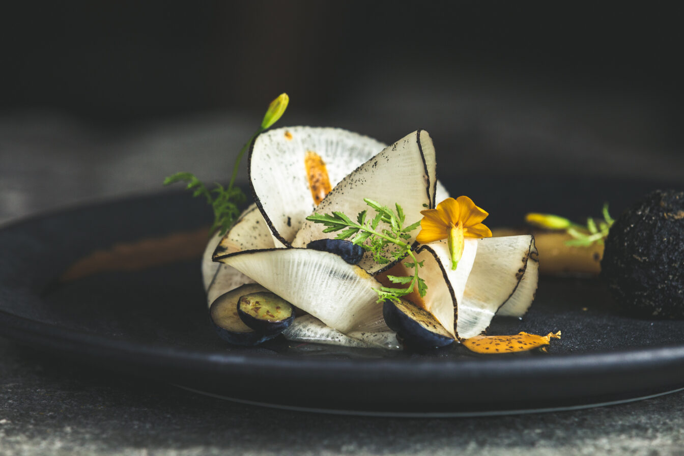 Gourmetgericht im Restaurant Kräuterreich by Vitus Winkler: kunstvoll angerichtete Komposition aus hauchdünnem Gemüse, Wildkräutern, Blüten und feinen Texturen auf dunklem Teller – moderne Alpenküche auf Sterne-Niveau.