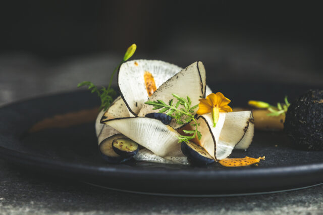 Gourmetgericht im Restaurant Kräuterreich by Vitus Winkler: kunstvoll angerichtete Komposition aus hauchdünnem Gemüse, Wildkräutern, Blüten und feinen Texturen auf dunklem Teller – moderne Alpenküche auf Sterne-Niveau.