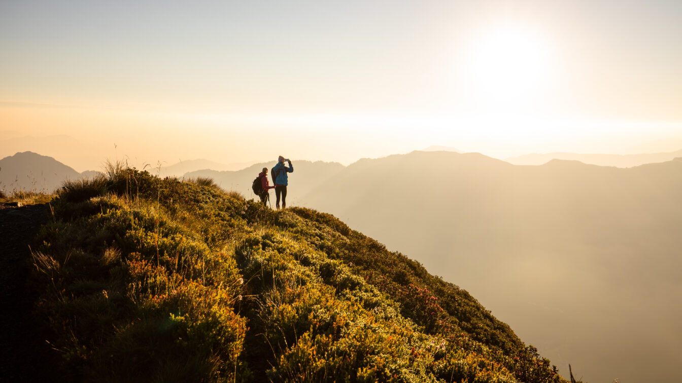 Sonnenaufgangswanderung am Wiedersbergerhorn bei Alpbach – zwei Wanderer genießen die goldene Morgenstimmung über dem Alpbachtal in Tirol, mit Blick auf die Bergsilhouetten im Gegenlicht.