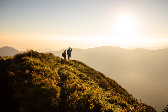 Sonnenaufgangswanderung am Wiedersbergerhorn bei Alpbach – zwei Wanderer genießen die goldene Morgenstimmung über dem Alpbachtal in Tirol, mit Blick auf die Bergsilhouetten im Gegenlicht.