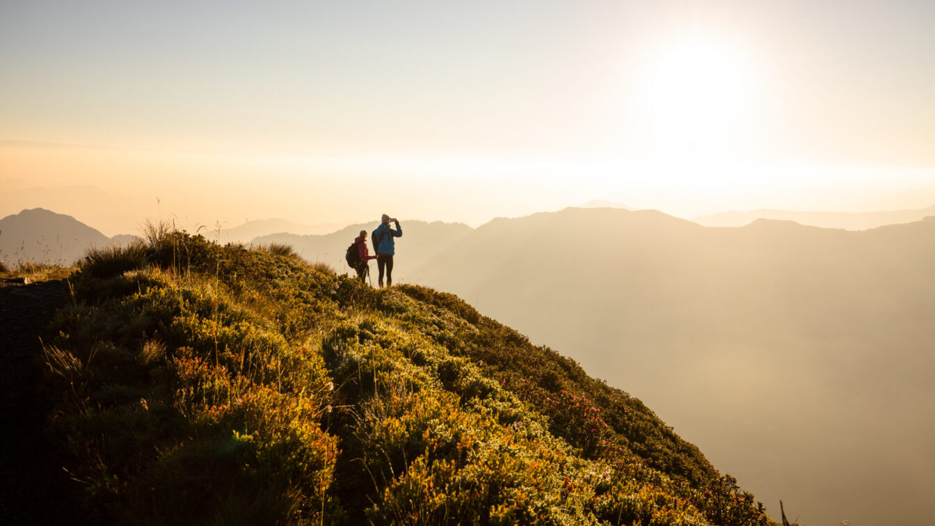 Sonnenaufgangswanderung am Wiedersbergerhorn bei Alpbach – zwei Wanderer genießen die goldene Morgenstimmung über dem Alpbachtal in Tirol, mit Blick auf die Bergsilhouetten im Gegenlicht.