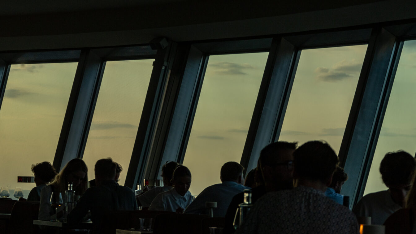 Stimmungsvolle Szene beim Sonnenuntergang über Berlin: Gäste sitzen in einem Restaurant mit großen Panoramafenstern und genießen den Ausblick auf den Himmel in warmem Abendlicht. Silhouetten der Menschen heben sich gegen den goldfarbenen Horizont ab – perfekter Moment für ein romantisches Dinner mit Blick auf den Sonnenuntergang in Berlin