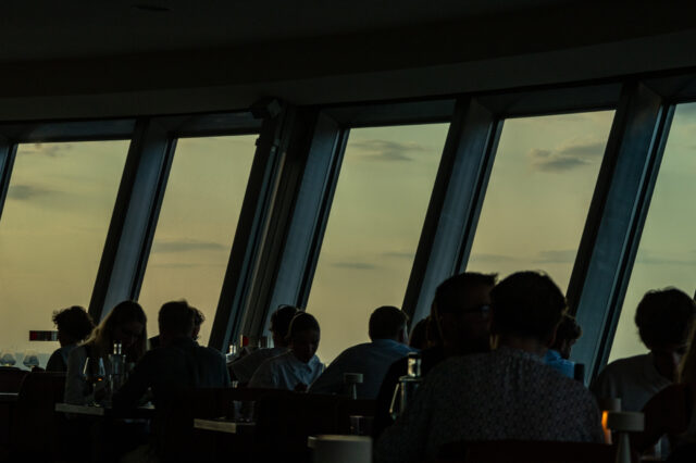Stimmungsvolle Szene beim Sonnenuntergang über Berlin: Gäste sitzen in einem Restaurant mit großen Panoramafenstern und genießen den Ausblick auf den Himmel in warmem Abendlicht. Silhouetten der Menschen heben sich gegen den goldfarbenen Horizont ab – perfekter Moment für ein romantisches Dinner mit Blick auf den Sonnenuntergang in Berlin