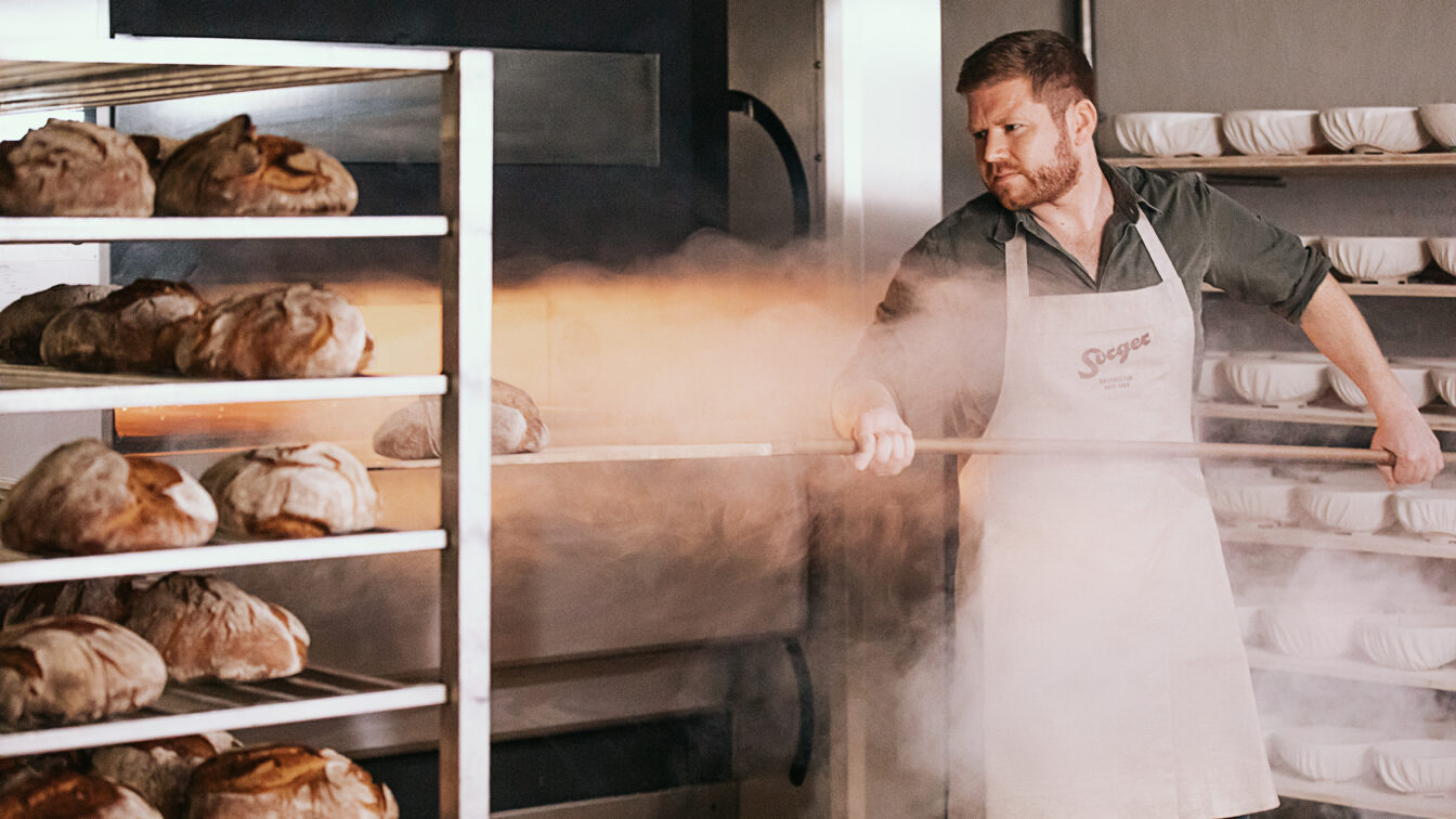 Ein Bäcker in Schürze mit Sorger-Logo zieht mit einem Holzschieber frisches Brot aus einem dampfenden Ofen. Im Hintergrund stehen Gärkörbchen ordentlich gestapelt in Regalen, daneben ein Metallwagen mit gebackenen Broten.
