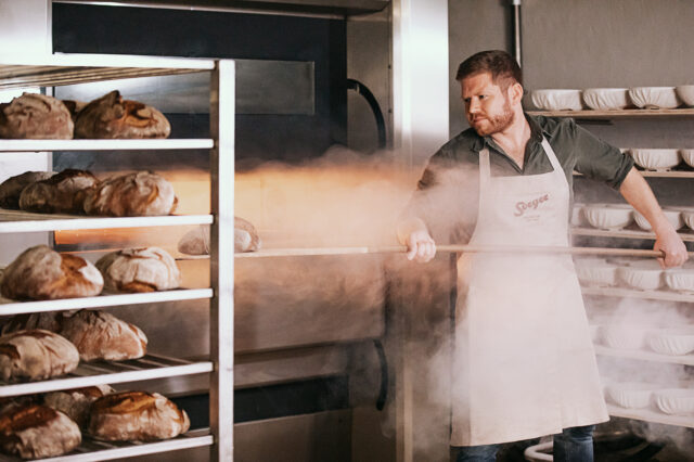 Ein Bäcker in Schürze mit Sorger-Logo zieht mit einem Holzschieber frisches Brot aus einem dampfenden Ofen. Im Hintergrund stehen Gärkörbchen ordentlich gestapelt in Regalen, daneben ein Metallwagen mit gebackenen Broten.