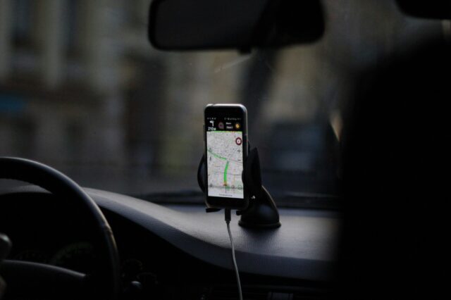 Ein Smartphone in einer Halterung auf dem Armaturenbrett eines Autos zeigt eine Navigations-App mit einer geplanten Route an. Das Auto befindet sich vermutlich im Stadtverkehr, Blickrichtung durch die Frontscheibe