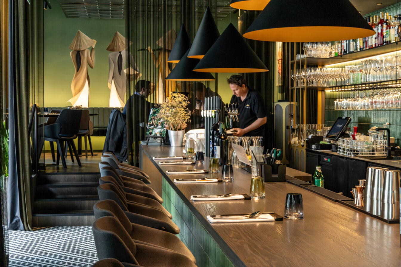 Stilvoller Barbereich im Vina Graz mit dunklem Holztresen, moderner Hängelampe und kunstvoller Wandmalerei von drei Figuren in vietnamesischer Tracht.