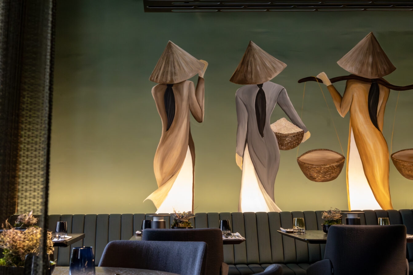 Künstlerisches Wandbild mit drei stilisierten Figuren in traditionellen vietnamesischen Hüten, die Körbe tragen – gemütlich eingerichtete Sitzecke im Vina Graz.