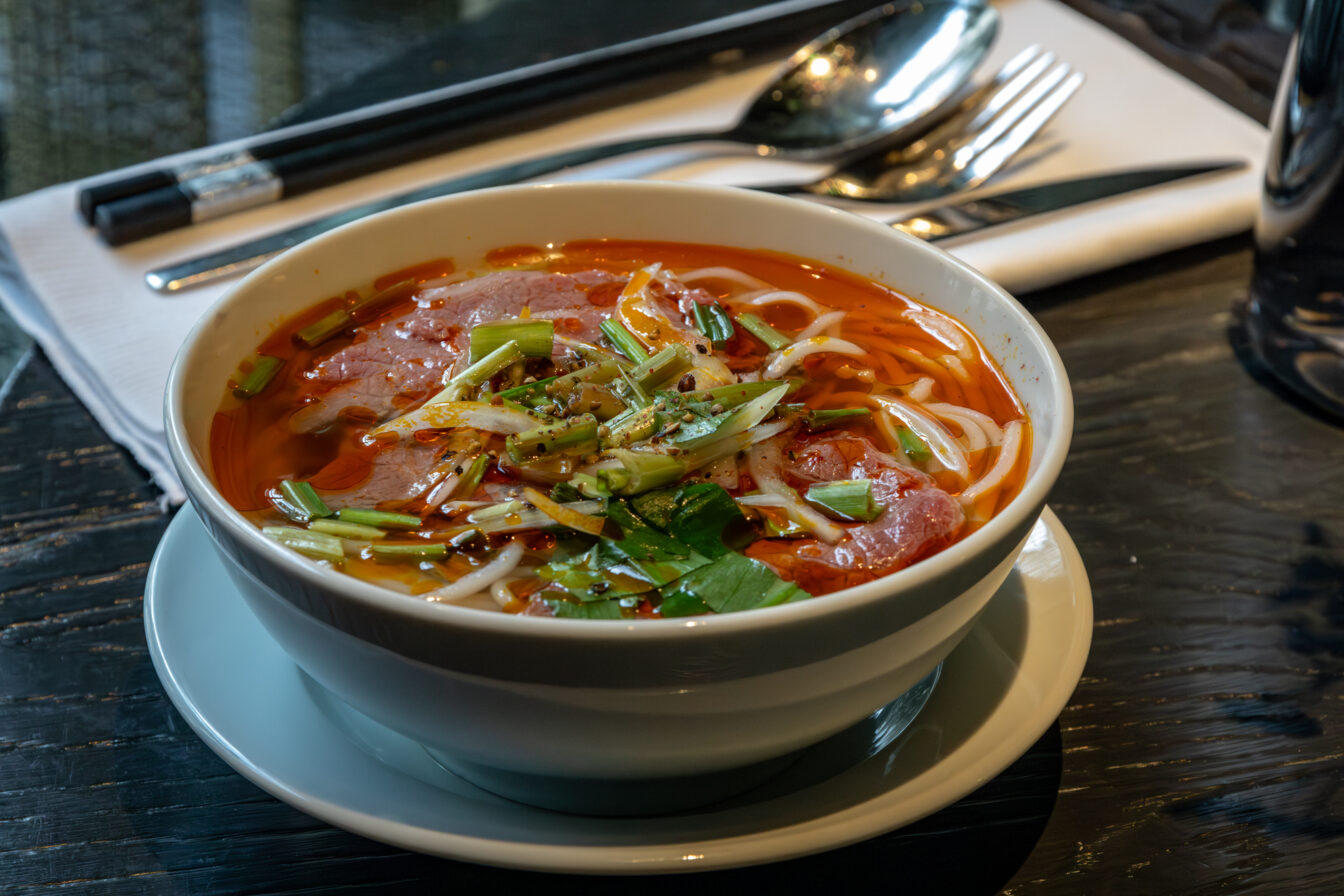 Nahaufnahme einer dampfenden vietnamesischen Nudelsuppe mit Rindfleisch, Frühlingszwiebeln und Kräutern auf einem gedeckten Tisch.