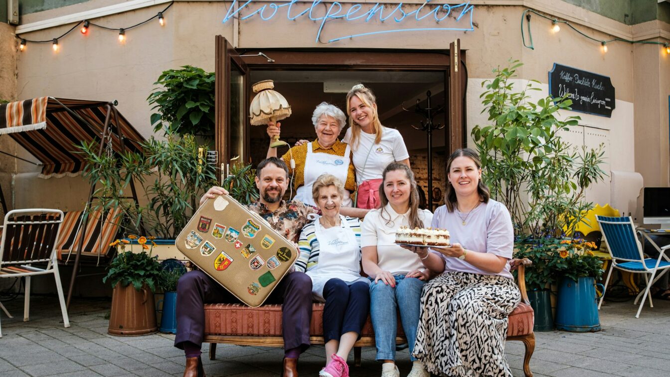 Sechs Personen vor einem Café in Wien, mit Kuchen und Koffer in der Hand.