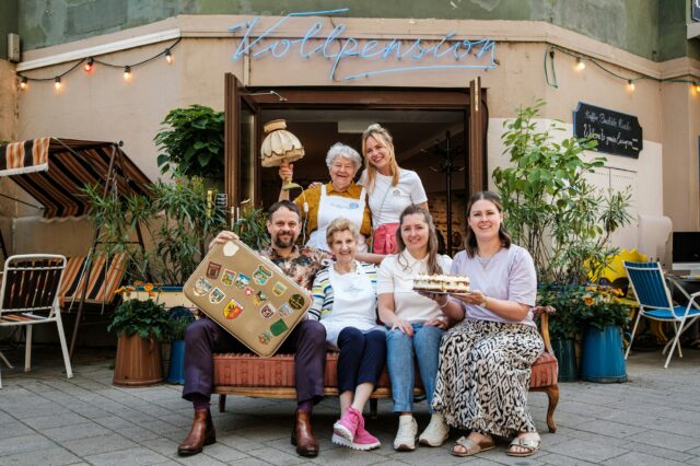 Sechs Personen vor einem Café in Wien, mit Kuchen und Koffer in der Hand.