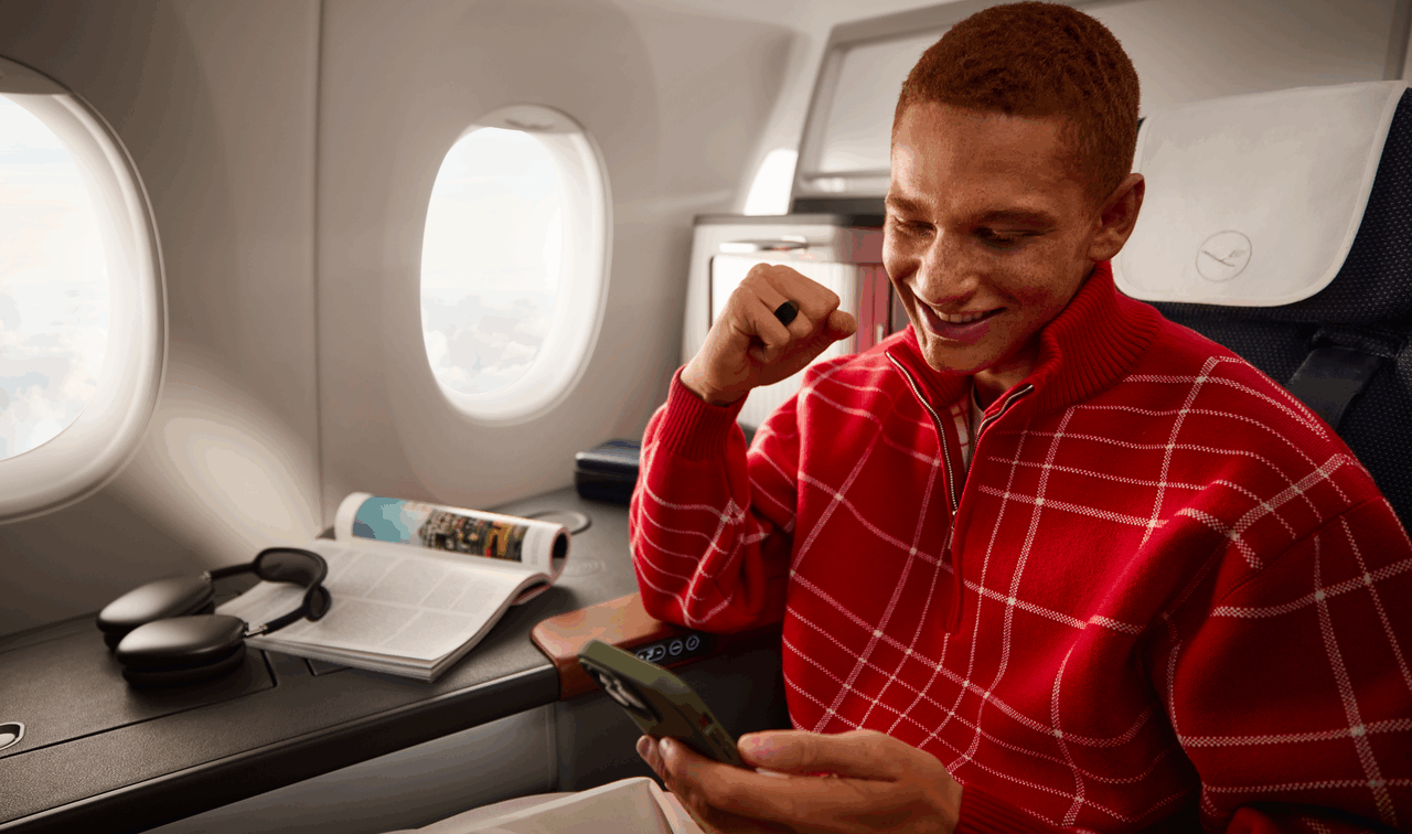 Ein junger Mann in rotem Pullover, der in der First Class eines Lufthansa Flugzeugs sitzt.