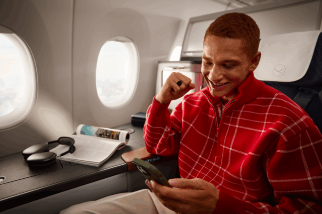 Ein junger Mann in rotem Pullover, der in der First Class eines Lufthansa Flugzeugs sitzt.