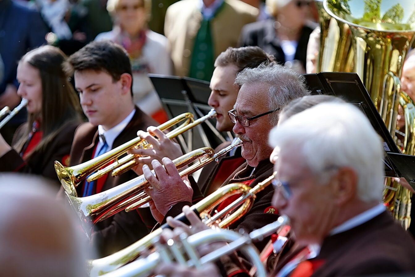 Blasmusikkapelle bei einem Promenadenkonzert im Freien – Musikerinnen und Musiker in traditioneller Tracht spielen Trompeten und Posaunen, Publikum im Hintergrund genießt die Darbietung.