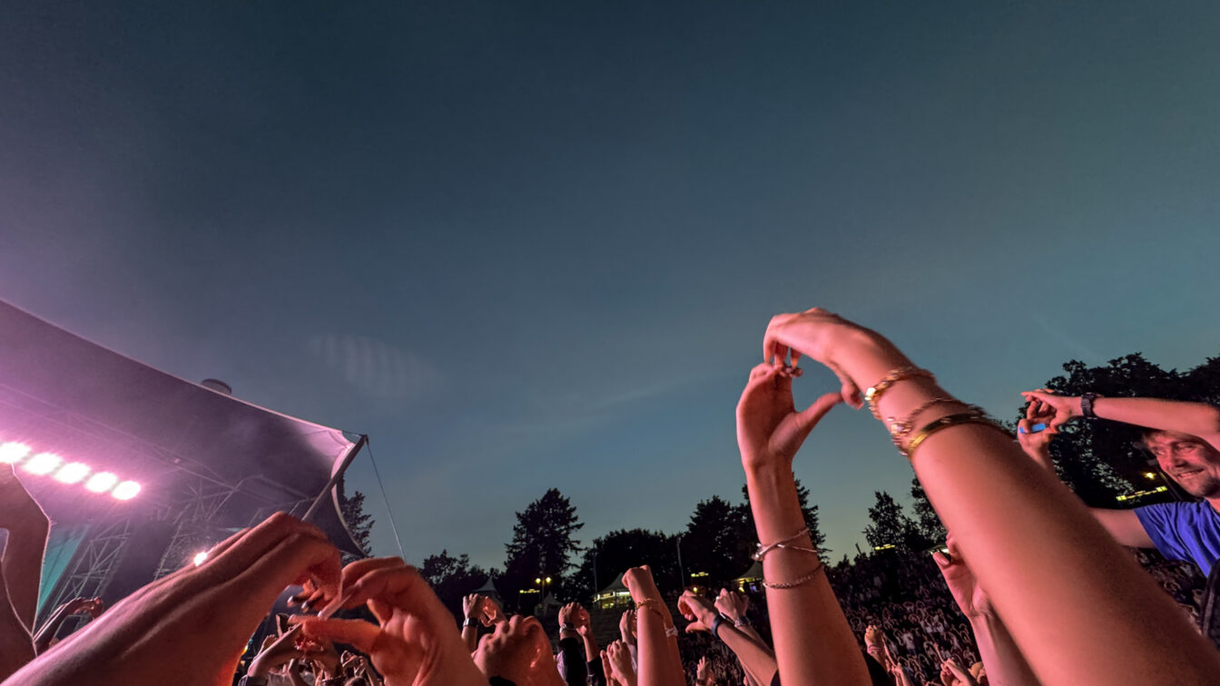 Ausgelassene Festivalstimmung beim Berlin Festival Sommer 2025: Zahlreiche Menschen stehen vor einer Bühne unter freiem Himmel in der Abenddämmerung, halten Hände in die Luft und formen Herzzeichen. Die Bühne ist in pink-violettes Licht getaucht, umgeben von Bäumen und sommerlicher Atmosphäre.