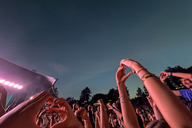 Ausgelassene Festivalstimmung beim Berlin Festival Sommer 2025: Zahlreiche Menschen stehen vor einer Bühne unter freiem Himmel in der Abenddämmerung, halten Hände in die Luft und formen Herzzeichen. Die Bühne ist in pink-violettes Licht getaucht, umgeben von Bäumen und sommerlicher Atmosphäre.