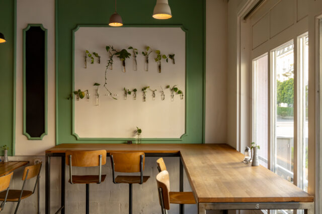Gemütlicher Innenraum eines modernen Cafés in Bern mit Holz- und Industriemöbeln, minimalistischer Pflanzen-Deko, großer Fensterfront und entspannter Atmosphäre – ideal für Frühstück, Lunch oder Kaffee in einem der stylischen Restaurants und Bars der Stadt.