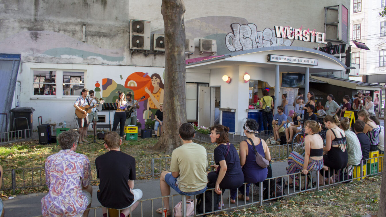 Eine Gruppe Menschen vor und um einen Würstelstand in Wien herum, die einem Konzert lauschen.