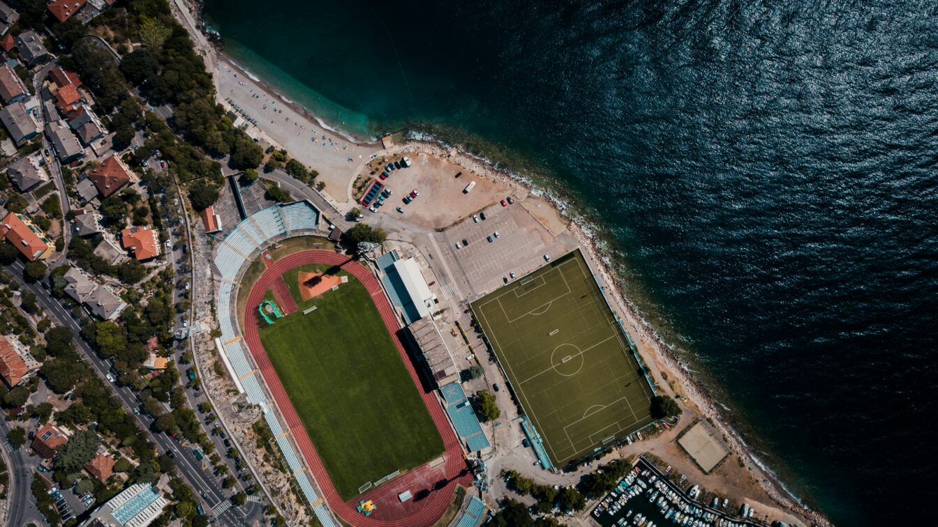 Eine Stadt am Meer mit Fußballstadion und Sportplatz aus der Vogelperspektive.