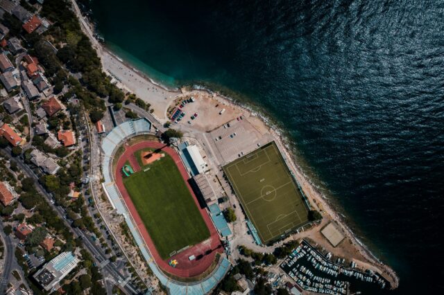 Eine Stadt am Meer mit Fußballstadion und Sportplatz aus der Vogelperspektive.