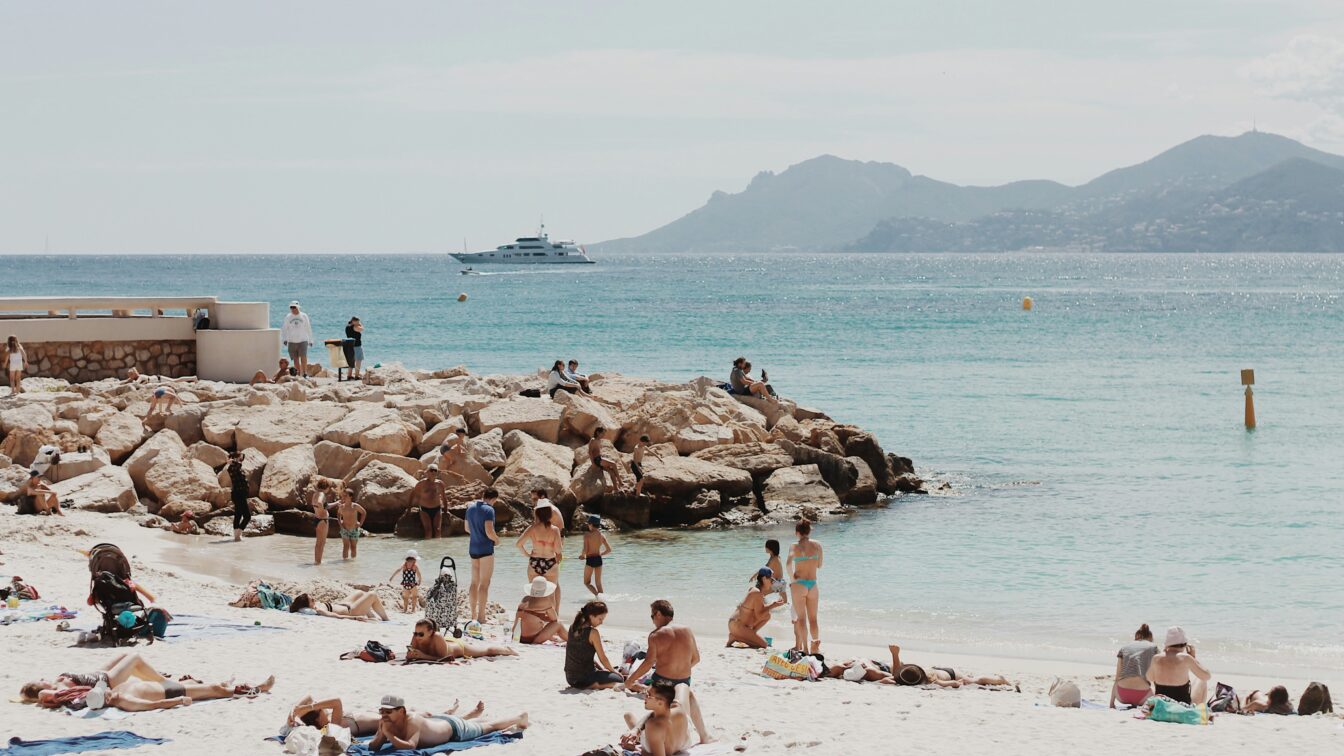 Menschen genießen einen sonnigen Tag an einem belebten Sandstrand in Südfrankreich. Einige liegen auf Handtüchern, andere baden im klaren türkisblauen Meer oder sitzen auf großen Felsen. Im Hintergrund ist ein luxuriöses weißes Schiff auf dem Wasser zu sehen, dahinter eine hügelige Küstenlandschaft. Die Atmosphäre wirkt entspannt und sommerlich.