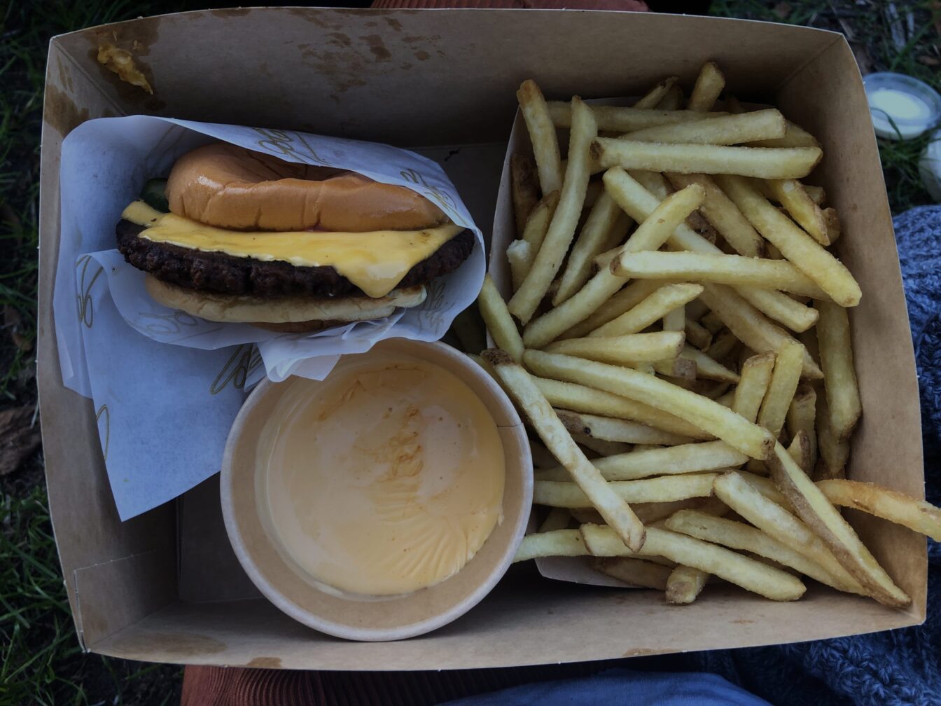 Burger-Menü von Goldies Berlin: In einer braunen Pappbox liegen knusprige Pommes, ein Cheeseburger mit fluffigem Bun und geschmolzenem Käse sowie ein Becher mit cremiger Käsesauce. Das Essen befindet sich auf einem Schoß im Freien, im Hintergrund ist etwas Gras zu sehen.